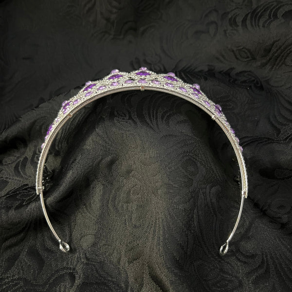 A silver-toned metal tiara or headband decorated with small clear rhinestones and clusters of purple stones, photographed on a dark textured fabric background.