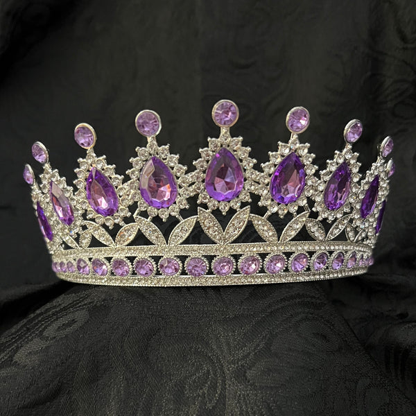 A silver tiara crown embellished with purple teardrop and round crystals, displayed on a textured black fabric background.