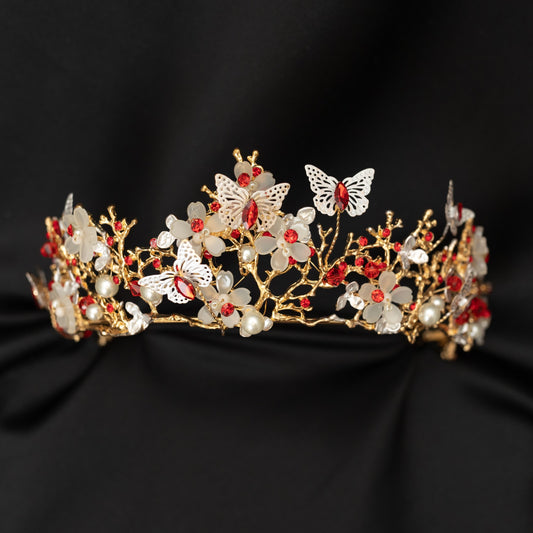 A gold-colored metal tiara adorned with white butterflies, leaf and flower designs, faux ruby red crystals, and faux pearl details on a black background. Front view 