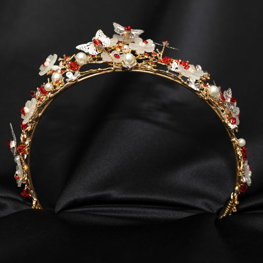 A gold-colored metal tiara adorned with white butterflies, leaf and flower designs, faux ruby red crystals, and faux pearl details on a black background. Bottom view 