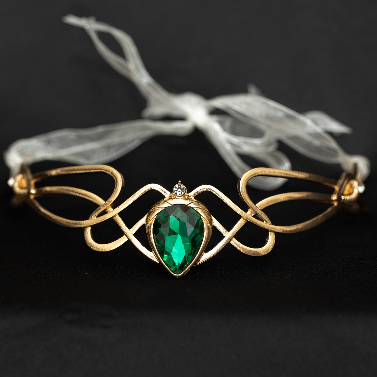 A gold-toned metal cuff bracelet featuring an intricate looped design and a central faceted green teardrop gemstone, displayed on a black background with a white ribbon tag.