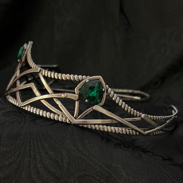 A silver-toned geometric cage-style bracelet with three green gemstones, displayed on a textured black fabric background.