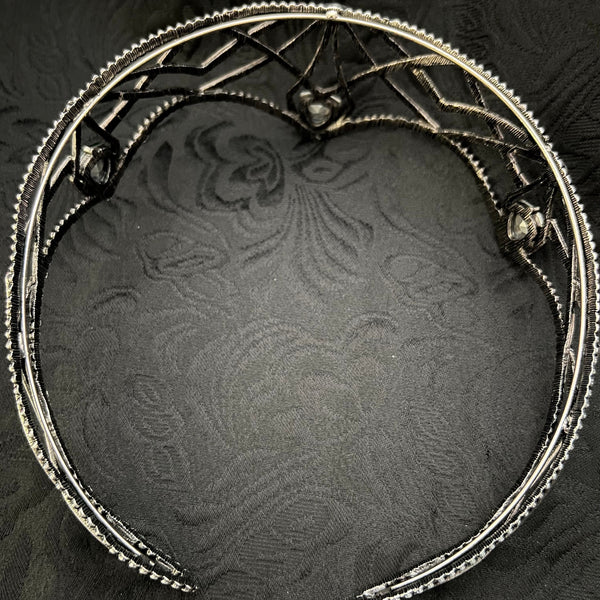 A silver-toned metal headband with a decorative filigree design and small spider motifs, displayed on a dark textured fabric background.