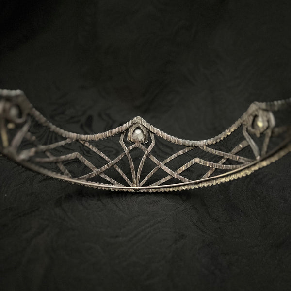 An ornate silver filigree tiara with geometric patterns and small pearl-like accents, displayed against a dark textured fabric background.