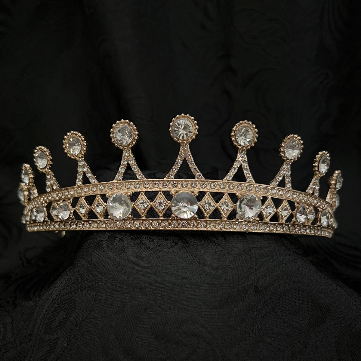 A gold-toned jeweled crown with clear gemstones, displayed against a dark black damask fabric background.