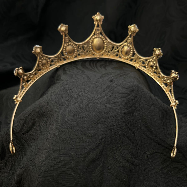 An ornate gold crown headband with intricate filigree detailing, displayed on a textured black fabric background.