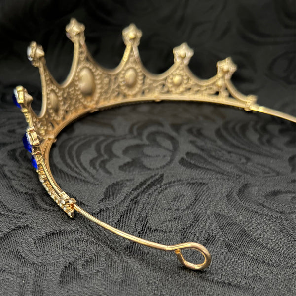 An antique gold crown-shaped brooch with a loop at the back and small blue gemstone accents, displayed on a textured black damask fabric background.