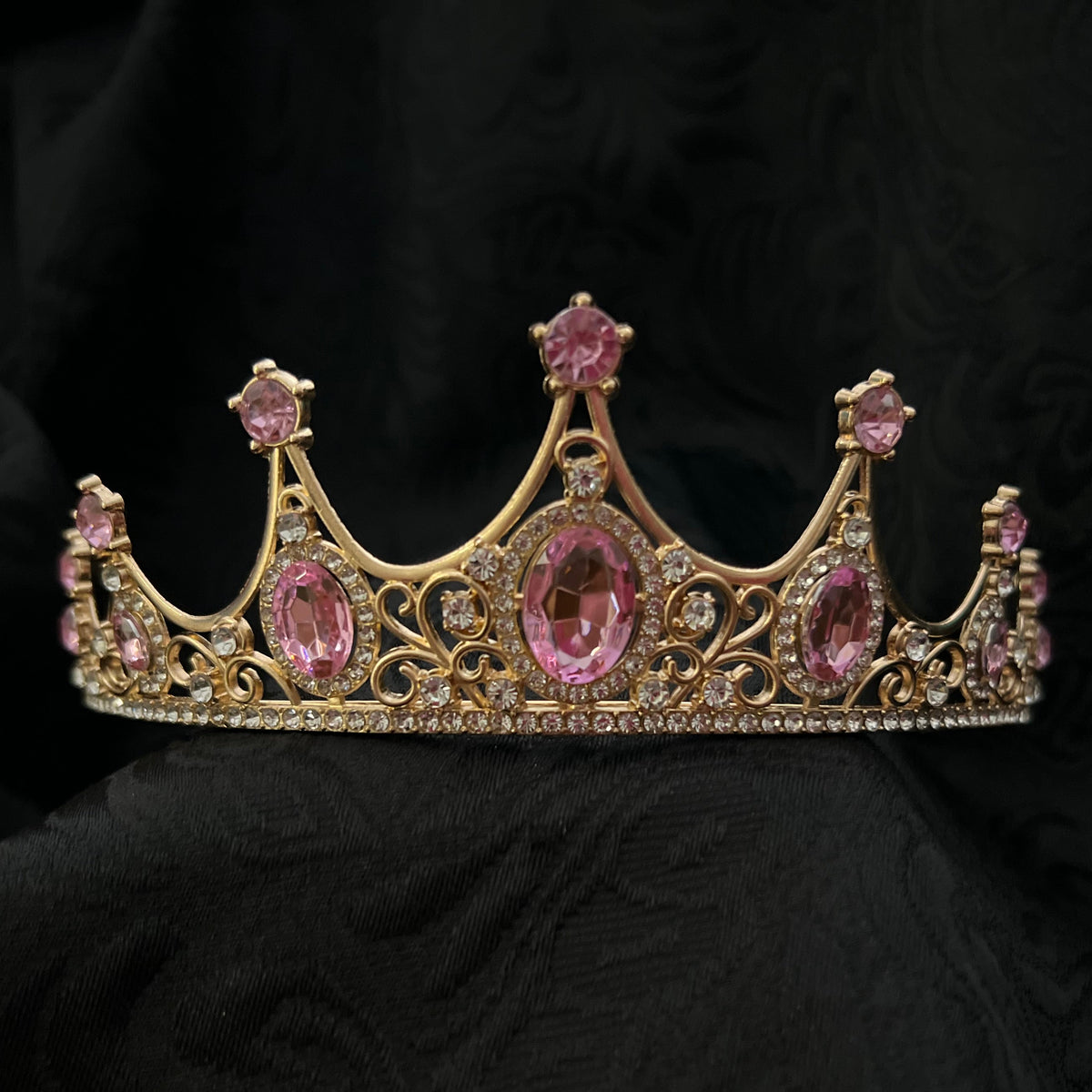 A gold-toned tiara crown adorned with pink gemstones, displayed on a draped black fabric background.