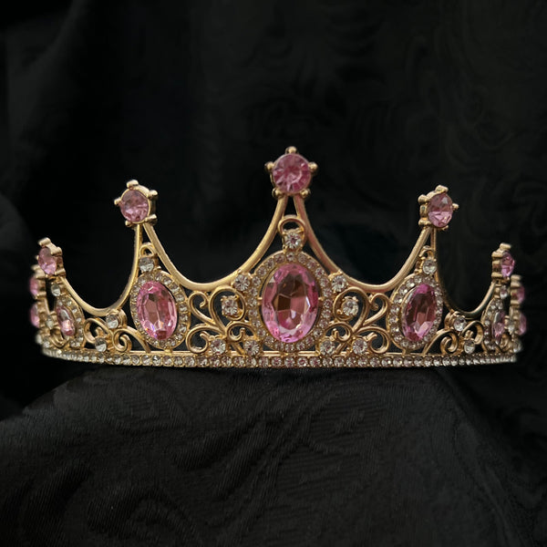 A gold-toned tiara crown adorned with pink gemstones, displayed on a draped black fabric background.