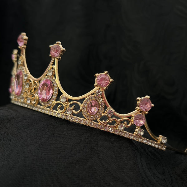 A gold tiara crown adorned with pink gemstones and small clear rhinestones, displayed on a textured black fabric background.r 