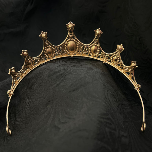 An ornate gold crown tiara with filigree detailing and central oval medallions, displayed on a draped black fabric background.