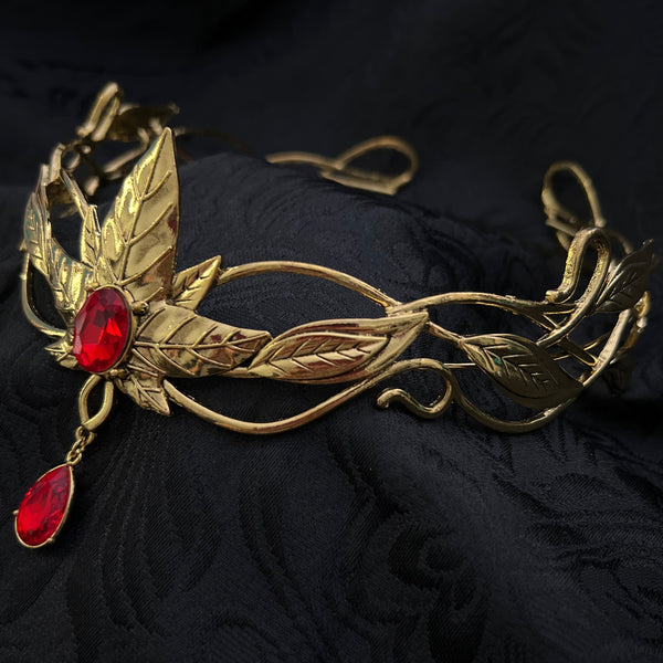 An ornate gold-toned metal brooch or pendant with leaf and vine motifs, accented by two red teardrop gemstones, displayed on a textured black fabric background.