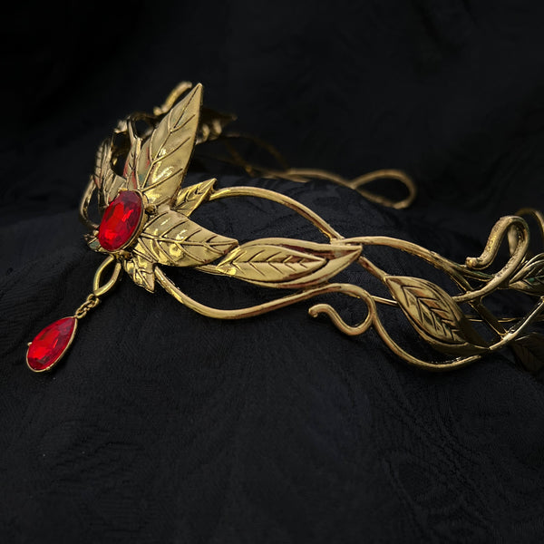 An ornate gold-toned metal headpiece or tiara featuring leaf and vine motifs, with two red teardrop gemstones, displayed on a textured black fabric background.