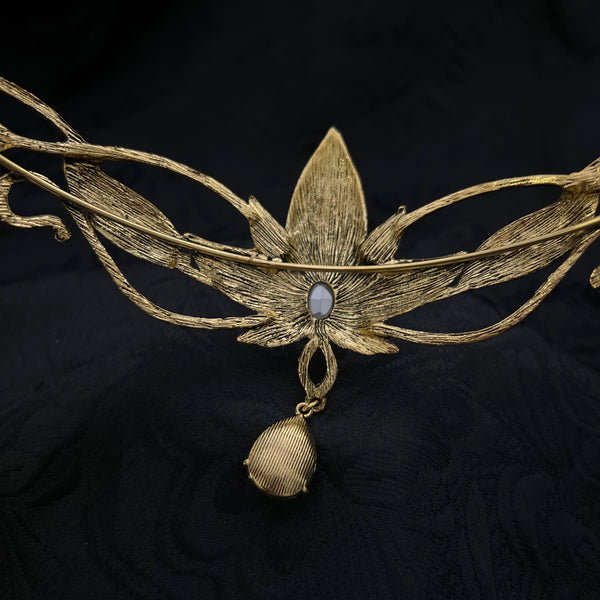An ornate gold-toned floral brooch or hair accessory with a small oval pale blue stone at the center, displayed on a textured black fabric background.
