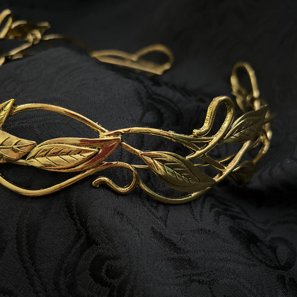 A close-up of a gold-toned metal vine or leaf ornament with detailed leaf and stem motifs, set against a dark black textured fabric background.