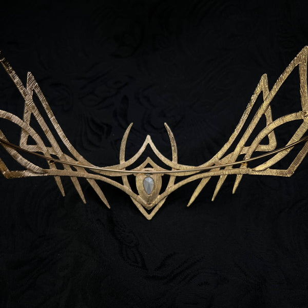 A gold-toned abstract metal wall sculpture with a central oval light blue stone, set against a textured black fabric background.