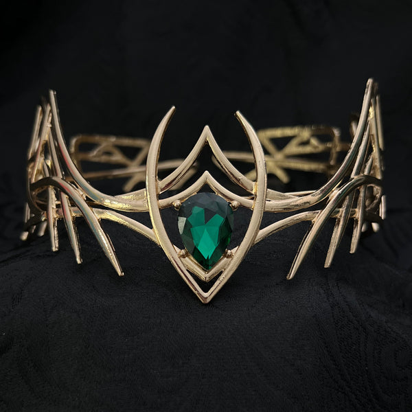 A gold-toned metal cuff bracelet featuring an intricate thorny eye design and a central green gemstone, displayed on a textured black fabric background.