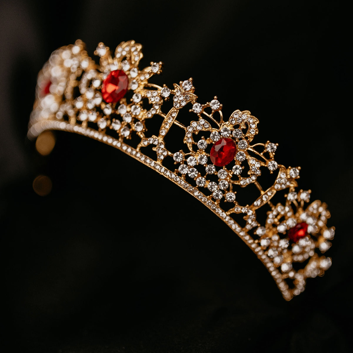 Benjamina's Tiara - A gold-colored metal tiara with clear crystal detailing and red gemstones set in the center, on a black background - Front Angle View 