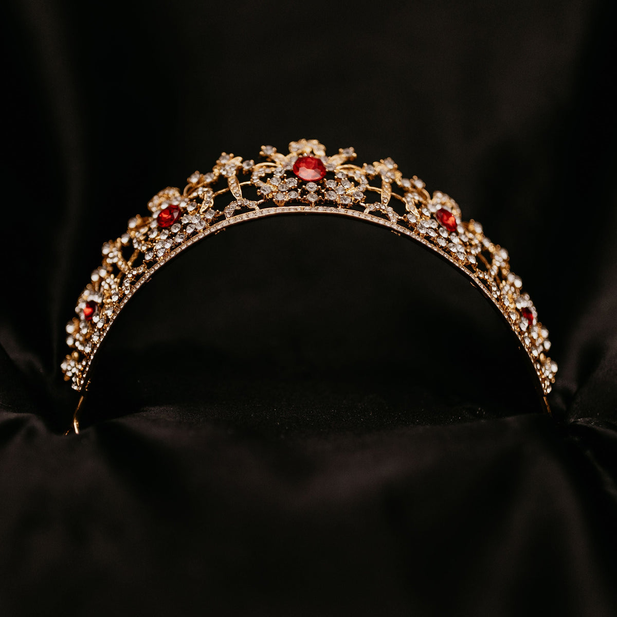 Benjamina's Tiara - A gold-colored metal tiara with clear crystal detailing and red gemstones set in the center, on a black background - Bottom View 