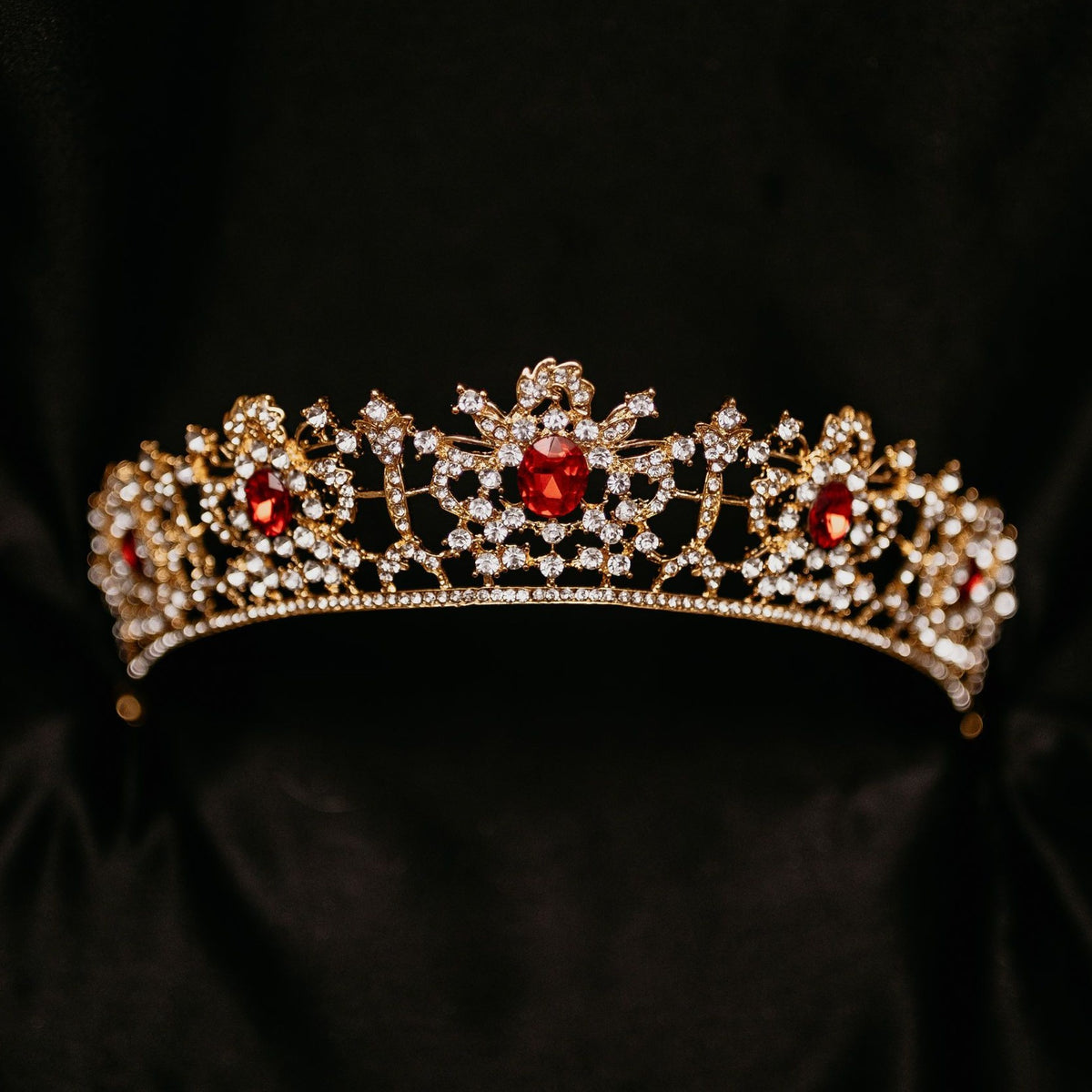 Benjamina's Tiara - A gold-colored metal tiara with clear crystal detailing and red gemstones set in the center, on a black background - front View 