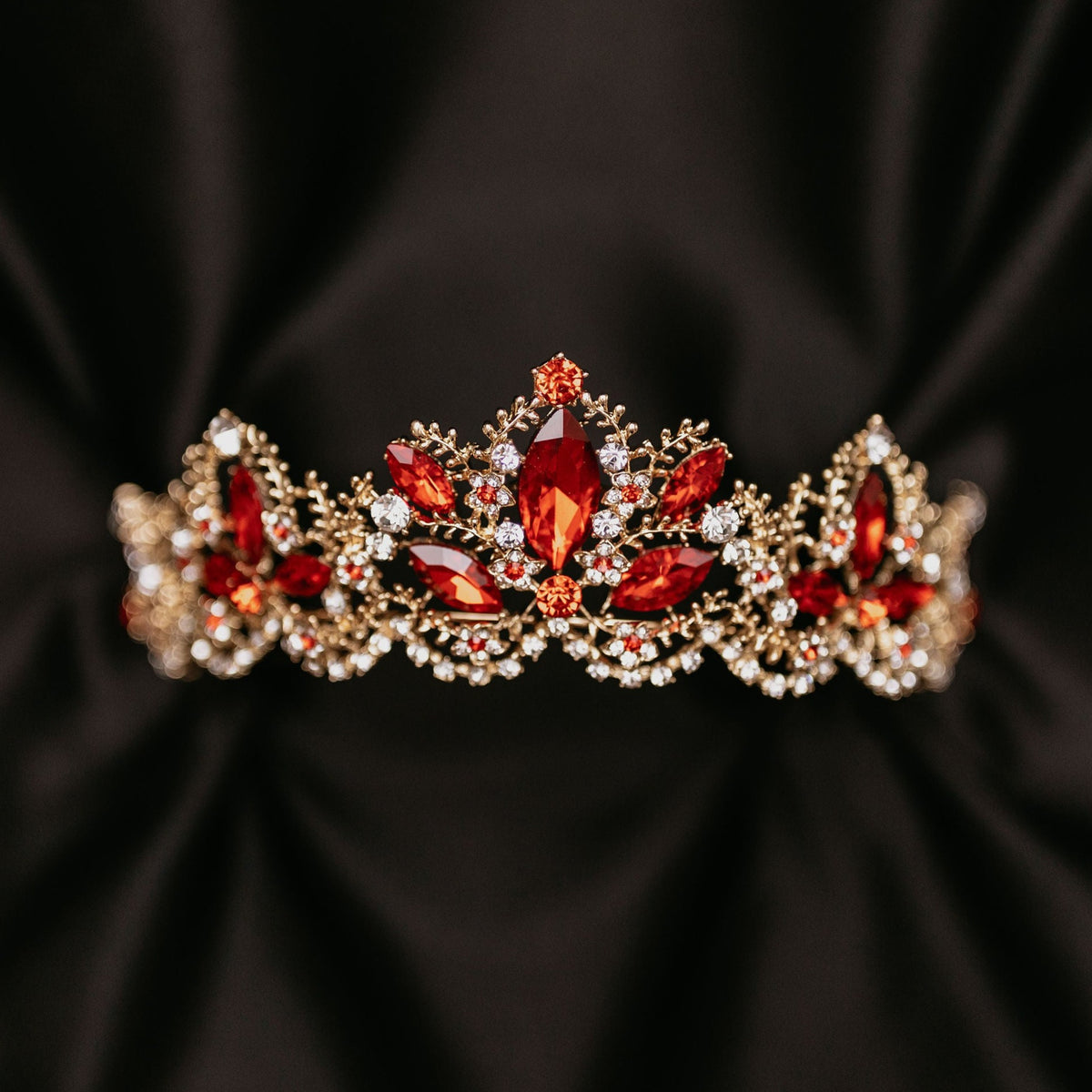 A red and gold tiara with intricate detailing and clear crystals, displayed against a black background.