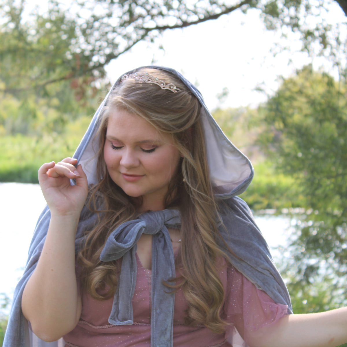 Elegant minimalist gold tiara with delicate crystal accents and classic curved design - Modeled by young woman with long blond hair and blue velvet hooded cape